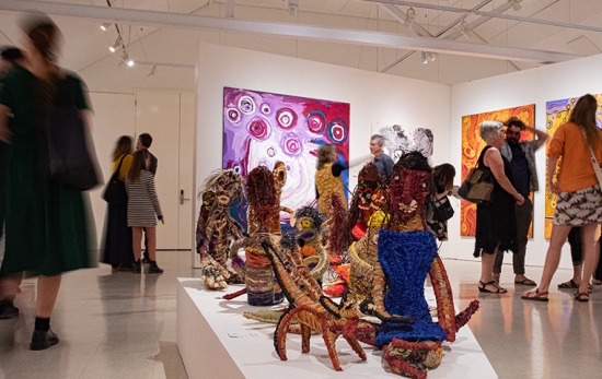 crowds walk through busy gallery, foreground shows woven sculptures of goannas and women sitting