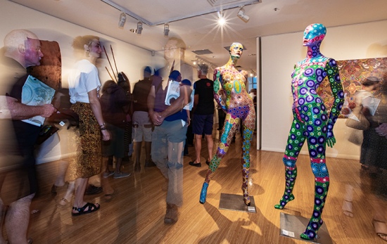crowds walk through busy gallery, foreground shows two mannequins painted in an aboriginal dot painting style